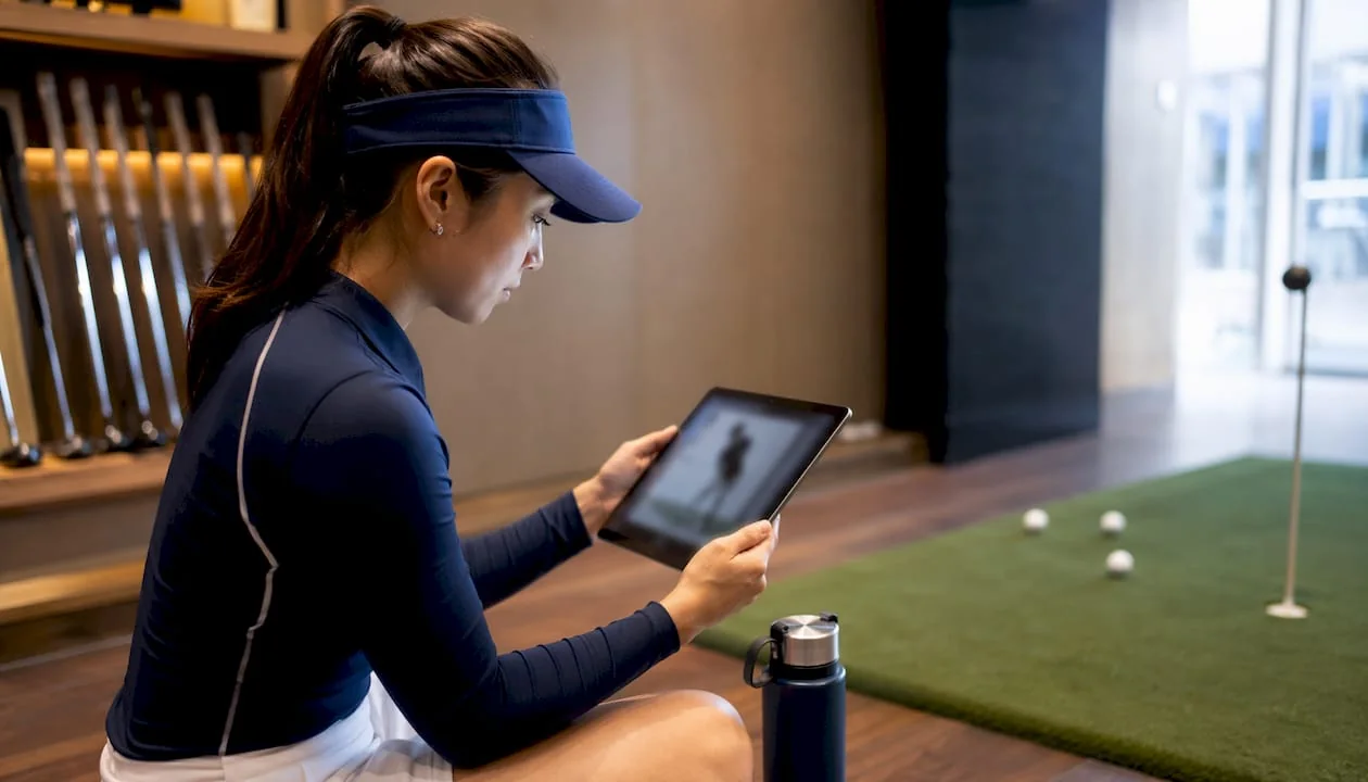 Golfer sitting indoors at a practice bay reviewing swing video on a tablet